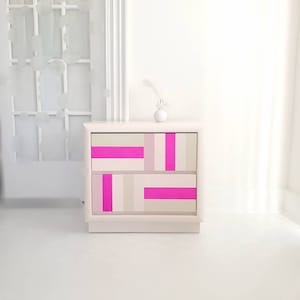 May include: A white nightstand with two drawers. The drawers are painted with a geometric pattern of pink and beige stripes.