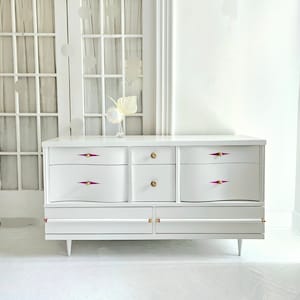 May include: White dresser with six drawers and gold hardware. The drawers have pink accents on the handles. The dresser is on a white floor in front of a white door.