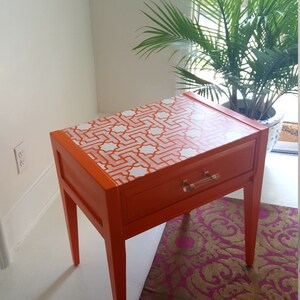 May include: Orange nightstand with a single drawer and white geometric pattern on the top. The nightstand has four legs and is sitting on a pink and gold patterned rug.