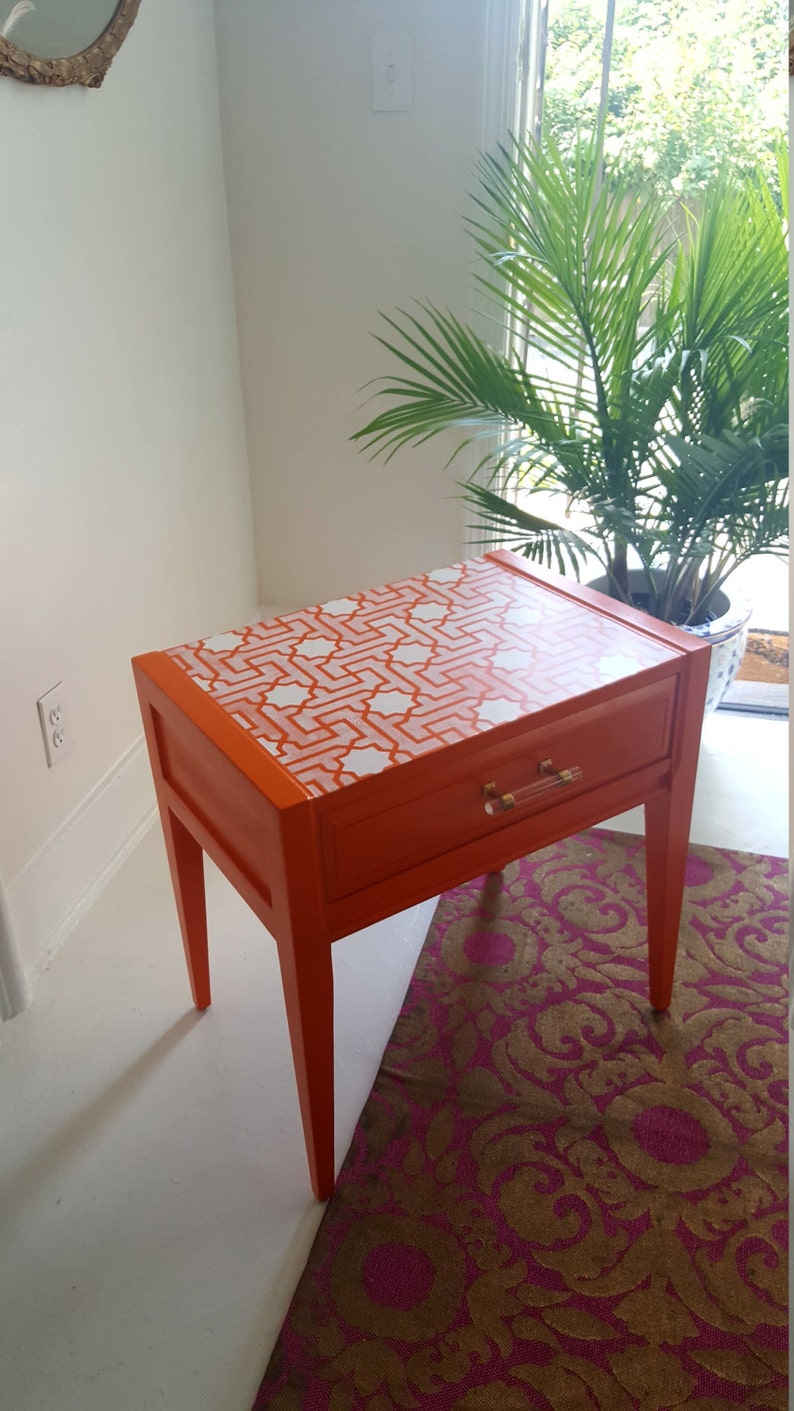 May include: Orange nightstand with a single drawer and white geometric pattern on the top. The nightstand has four legs and is sitting on a pink and gold patterned rug.