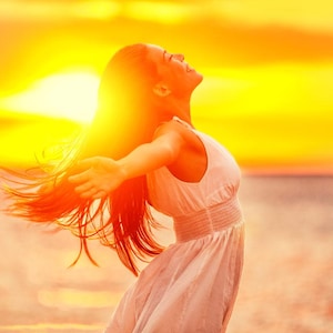 Pode incluir: Uma mulher com cabelo comprido e ruivo está de pé com os braços estendidos, de frente para o pôr do sol. Ela está vestindo um vestido branco e tem uma expressão de alegria no rosto.