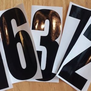 May include: Black vinyl number decals, including the numbers 1, 6, 3, and 2, are displayed on a light wood surface. The numbers are bold and glossy, with a white background. These decals are suitable for various DIY projects.