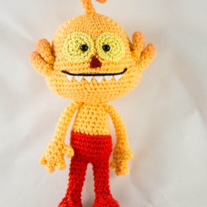 May include: A crocheted plush toy with a yellow body, red pants, and a toothy grin. The toy has large yellow eyes with black pupils and a red nose.