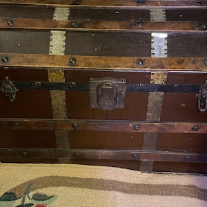 May include: A vintage wooden trunk with metal accents and hardware. The trunk has a brown finish and is decorated with brass and silver accents. The trunk has a hinged lid and two metal clasps.