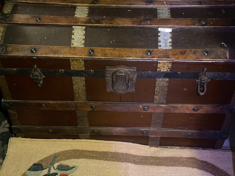 May include: A vintage wooden trunk with metal accents and hardware. The trunk has a brown finish and is decorated with brass and silver accents. The trunk has a hinged lid and two metal clasps.