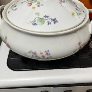 May include: White ceramic serving dish with lid, adorned with floral patterns in pink, purple, and yellow. The dish has two handles and a knob on the lid. It is sitting on a white stove top.
