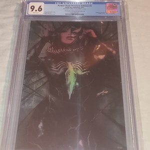 May include: A graded comic book in a protective case. The comic book cover features a black and green illustration of a woman with a symbiote costume. The comic book is graded 9.6 by CGC. The comic book is titled "Power Hour Preview Edition #2" and is published by Black Ops Publishing in 2023. The comic book is number 17 of 20.