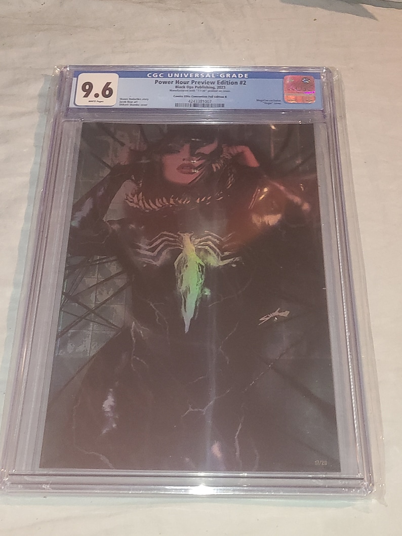 May include: A graded comic book in a protective case. The comic book cover features a black and green illustration of a woman with a symbiote costume. The comic book is graded 9.6 by CGC. The comic book is titled "Power Hour Preview Edition #2" and is published by Black Ops Publishing in 2023. The comic book is number 17 of 20.