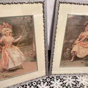 May include: Two framed vintage prints of women in antique clothing. The print on the left shows a woman in a white and pink dress on a swing. The print on the right shows a woman in a white and pink dress sitting on a rock.