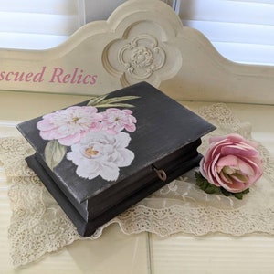 May include: A black wooden box with a hinged lid, decorated with a floral design featuring pink and white peonies. The box has a keyhole and is sitting on a white lace doily.