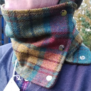 May include: A multi-coloured plaid neck warmer in teal, burgundy, and gold tweed fabric. It features silver snap closures. The neck warmer is designed to be worn around the neck.