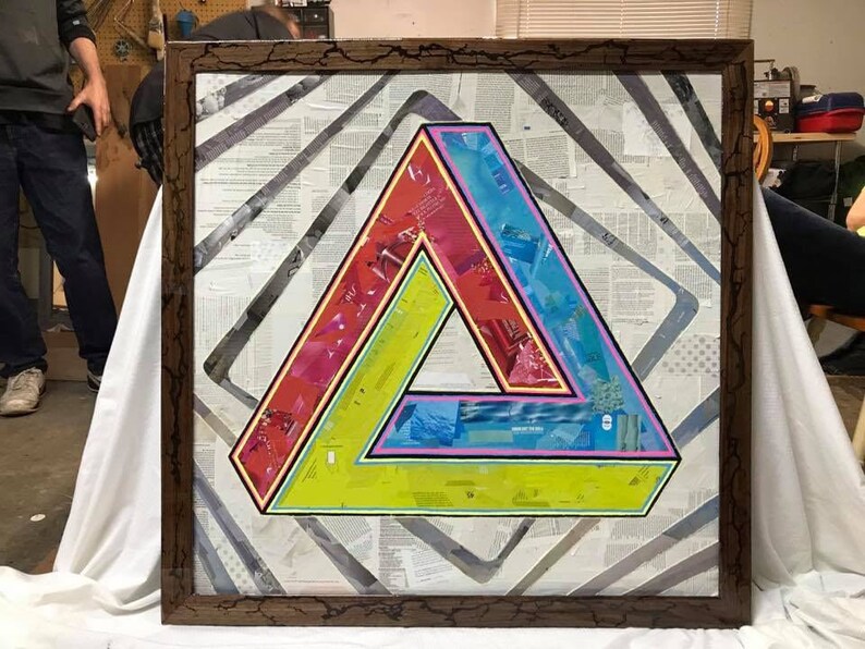 May include: A framed artwork featuring a colorful, optical illusion triangle design. The triangle is made up of overlapping layers of paper with different colors and patterns. The artwork is set against a background of white paper with a subtle gray pattern.