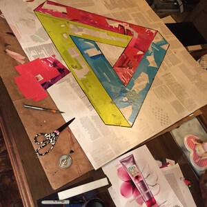 May include: A large, colorful paper art project depicting a Penrose triangle. The triangle is made of red, yellow, blue, and white paper. The project is in progress and is being worked on at a table.