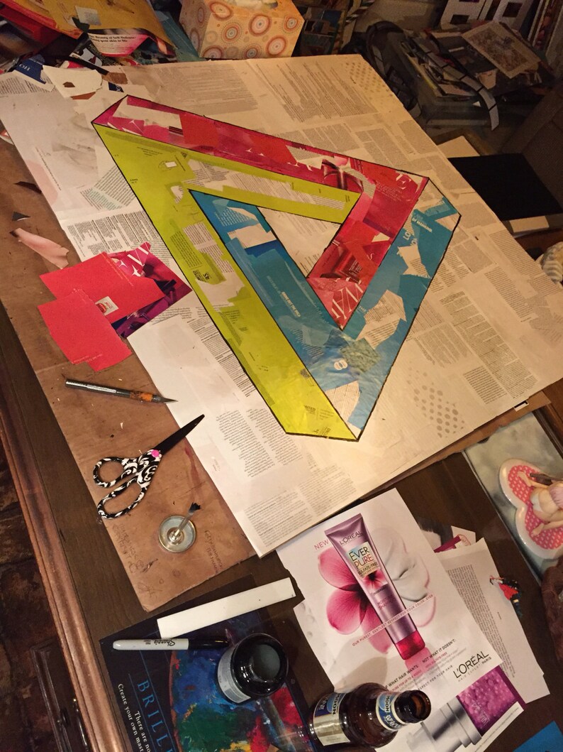 May include: A large, colorful paper art project depicting a Penrose triangle. The triangle is made of red, yellow, blue, and white paper. The project is in progress and is being worked on at a table.