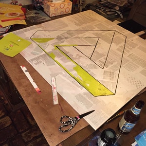 May include: A close-up of a table with a large sheet of paper with a black triangle drawn on it. The triangle is filled in with yellow paint. There are various art supplies on the table, including scissors, a ruler, a bottle of glue, and a bottle of blue liquid.