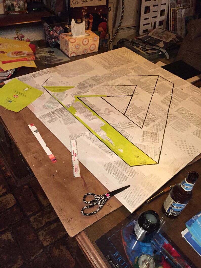 May include: A close-up of a table with a large sheet of paper with a black triangle drawn on it. The triangle is filled in with yellow paint. There are various art supplies on the table, including scissors, a ruler, a bottle of glue, and a bottle of blue liquid.
