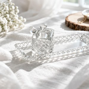 May include: A clear, cut-glass perfume bottle with a geometric design and a matching rectangular tray. The bottle has a faceted stopper. The items are displayed on a white, textured surface, with soft lighting.