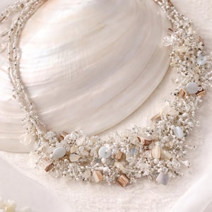 Op de afbeelding: Een delicate ketting met meerdere strengen kleine zilveren kralen, geaccentueerd met clusters van witte, beige en lichtblauwe kralen. De ketting wordt gepresenteerd op een grote, witte zeeschelp, wat een zachte, elegante uitstraling creëert.