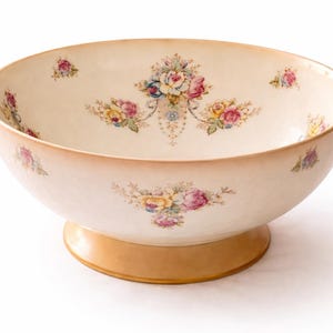 Könnte beinhalten: Cremefarbene Keramikschale mit Sockel, verziert mit Blumenmustern in Rosa, Gelb und Blau. Die Schale hat ein breites, flaches Design, ideal zum Servieren. Der Sockel ist in einem warmen, hellbraunen Farbton gehalten.