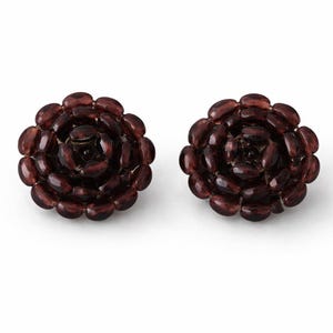 May include: A pair of dark red, flower-shaped earrings. Each earring is composed of numerous oval-shaped beads arranged in a spiral pattern, resembling a rose. The earrings are set against a white background.