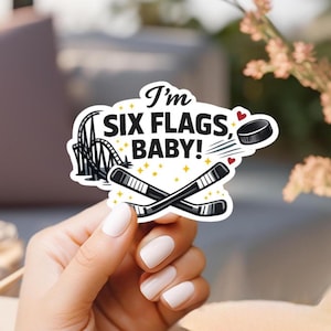 May include: A die-cut sticker featuring a black roller coaster silhouette, two crossed black hockey sticks, and a black hockey puck. The text reads I'm SIX FLAGS, BABY! in black capital letters, accented by small yellow stars and two red hearts.