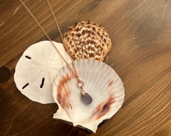 May include: A rose gold necklace with a dark gray sea glass pendant. The necklace is displayed on a seashell.