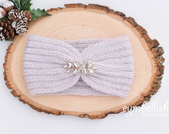 May include: A light purple knitted headband with a sparkly rhinestone embellishment. The headband is on a wooden surface.