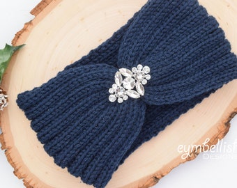 May include: Navy blue knitted headband with a large, clear rhinestone embellishment. The headband is twisted and has a knot in the center. The headband is on a wooden surface.
