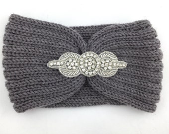 May include: A gray knitted headband with a decorative silver and rhinestone embellishment. The headband is a wide, ear-covering style, perfect for keeping warm. The embellishment is centered and adds a touch of sparkle.