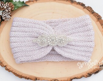 May include: A light gray knitted headband with a decorative silver embellishment. The headband features a twisted design and is adorned with a sparkling, circular rhinestone centerpiece. The accessory is displayed on a wooden surface.