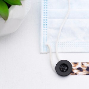 May include: A black button with a white string attached to a leopard print fabric mask extender.