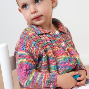 Può includere: Un bambino piccolo indossa un cardigan a righe colorate con colletto bianco e bottoni. Il cardigan è lavorato a maglia con una varietà di colori, tra cui rosa, blu, verde, giallo e arancione.