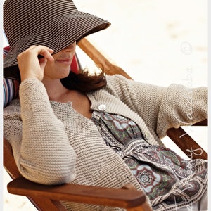 May include: A person wearing a beige knitted cardigan and a patterned dress, seated in a wooden beach chair. They are wearing a wide-brimmed striped hat, shielding their face. The scene suggests relaxation and leisure.