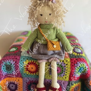 May include: A knitted doll with blonde curly hair wearing a green sweater, a purple skirt, and red shoes. The doll is sitting on a colourful crocheted blanket.