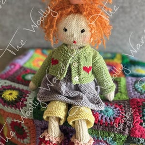 Może przedstawiać: Dzianinowa lalka z czerwonymi kręconymi włosami, ubrana w zielony kardigan z czerwonymi sercami, fioletową sukienkę, żółte spodnie i czerwone buty. Lalka siedzi na kolorowym szydełkowym kocu.