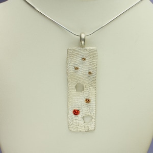 May include: A silver pendant necklace with a rectangular pendant featuring a textured surface and small red and orange gemstones.