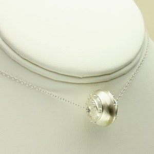 May include: A silver necklace with a textured, round pendant. The pendant is a cylinder shape with a slightly domed top.