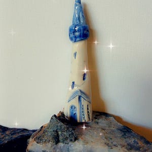 May include: A miniature ceramic lighthouse, predominantly white with blue detailing. The top and door are blue. The lighthouse is displayed on a rock, evoking a coastal scene. The height is approximately 10 cm.