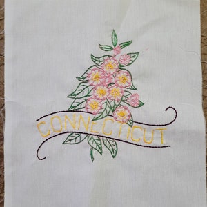 May include: Embroidered white fabric square featuring a floral design with pink and yellow flowers, green leaves, and the word "CONNECTICUT" in yellow script. The text is on a curved banner.
