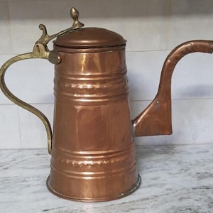 May include: A copper-colored vintage tea kettle with a lid and a curved spout. The kettle has a brass handle and accents. The design is cylindrical with horizontal ridges. The kettle is likely used for brewing tea or coffee.