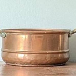 May include: A copper-colored, round pot with two small handles. The pot has a hammered texture and sits on a wooden surface. The pot is a kitchenware item.