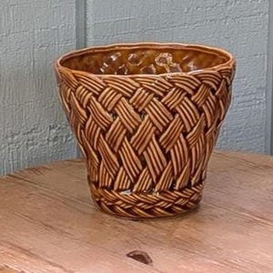 May include: A brown ceramic planter featuring a glossy finish and a textured exterior that mimics the appearance of woven basketry. The vessel has a wide, open circular top and a tapered base, showcasing a consistent brown glaze throughout.