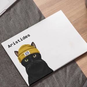 May include: A white notepad with the name "Aristides" printed in black. The notepad features a black cat wearing a yellow beanie with a blue and white design. The cat is looking at the viewer. The notepad is on a wooden surface.