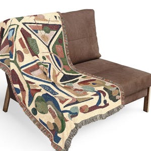 May include: A woven throw blanket featuring an off-white base with a multi-colored abstract geometric pattern. The design includes various shapes in shades of dark blue, forest green, terracotta, and tan, finished with a fringed edge detail.