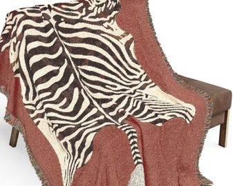 May include: A woven throw blanket with a zebra print design. The blanket features a rust-colored base with a zebra pattern in black and cream. The blanket has a fringed edge and is draped over a bench.