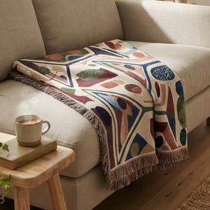May include: A woven throw blanket featuring a geometric pattern with shapes in shades of navy blue, forest green, terracotta, and cream. The blanket has a fringed edge and is draped over a light beige sofa, showcasing a textured fabric design.