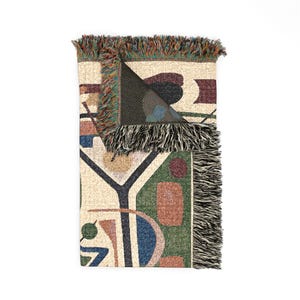 May include: A woven throw blanket featuring an abstract pattern of geometric shapes in shades of green, blue, red, and cream. The blanket has a folded top corner revealing a dark interior and is finished with multicolored fringe along the edges.
