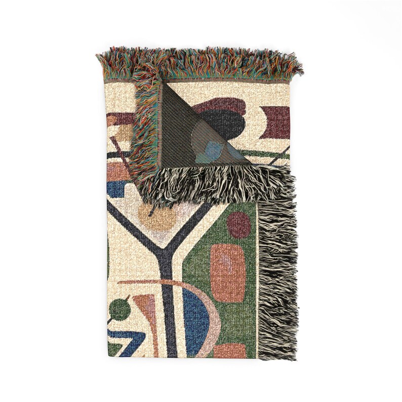 May include: A woven throw blanket featuring an abstract pattern of geometric shapes in shades of green, blue, red, and cream. The blanket has a folded top corner revealing a dark interior and is finished with multicolored fringe along the edges.