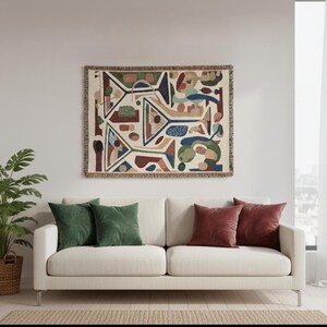 May include: A rectangular woven wall tapestry featuring an abstract pattern of geometric shapes and lines in shades of green, blue, red, and tan. The textile piece has a fringed border on all four sides and is displayed horizontally.