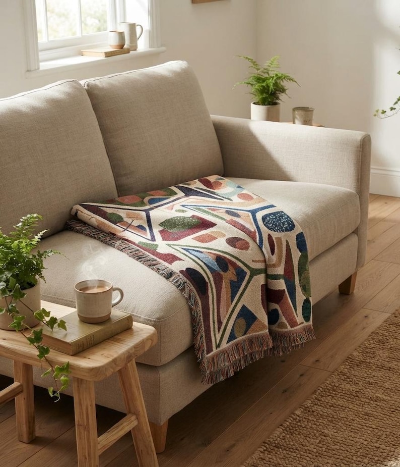 May include: A woven throw blanket featuring a geometric pattern with shapes in shades of navy blue, forest green, terracotta, and cream. The blanket has a fringed edge and is draped over a light beige sofa, showcasing a textured fabric design.